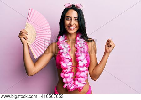 Beautiful hispanic woman wearing bikini holding handfan screaming proud, celebrating victory and success very excited with raised arm 