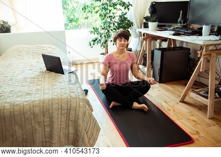 A Woman Sitting On A Mat In Front Of Bed While Doing Yoga Position In Bedroom. Relaxation Yoga Pract