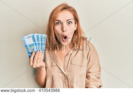 Young caucasian woman holding hungarian forint banknotes scared and amazed with open mouth for surprise, disbelief face 