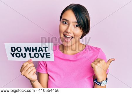 Beautiful young woman with short hair holding paper with do what you love text pointing thumb up to the side smiling happy with open mouth 