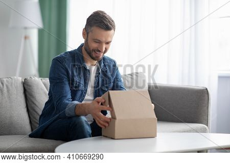 Happy Middle-aged Man Unpacking Delivery Box, Sitting On Couch By Tea Table, Home Interior. Satisfie