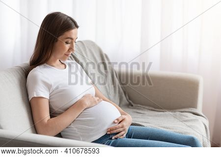 Prenatal Time. Beautiful Future Mother Sitting On Couch Embracing Her Belly, Happy Pregnant Woman Re