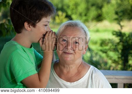 Boy whispering to grandma in summer outdoors garden, grandkid and grandmother lalking.