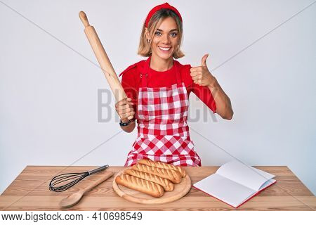 Beautiful caucasian woman wearing baker apron making homemade bread smiling happy and positive, thumb up doing excellent and approval sign 