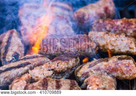 Preparing Meat Rolls Called Mici Or Mititei On Barbecue. Close Up Of Grill With Burning Fire With Fl
