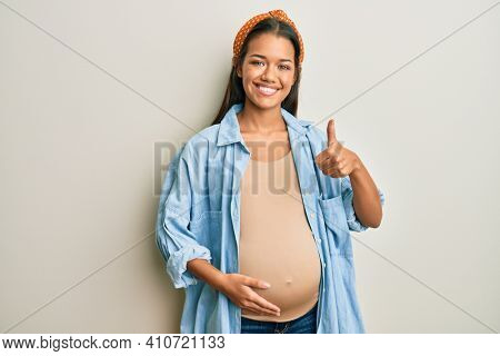 Beautiful hispanic woman expecting a baby, touching pregnant belly doing happy thumbs up gesture with hand. approving expression looking at the camera showing success. 