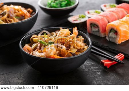 Traditional Asian food set. Wok noodles with shrimps and vegetables in a black bowl, sushi roll and seaweed salad on a black textured background, angle view.