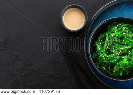 Seaweed salad with sesame seeds in a black bowl, peanut sauce and chopsticks on a black background with texture. View from above, copy space.