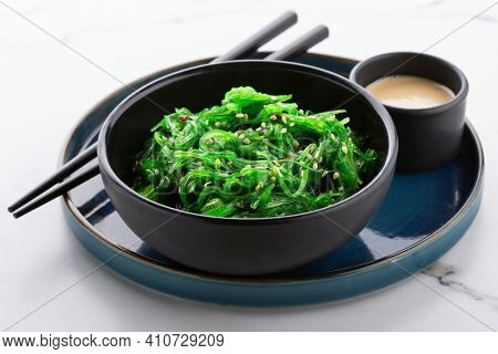 Healthy green seaweed salad with sesame seeds in a black bowl and peanut sauce on white stone background, angle view.