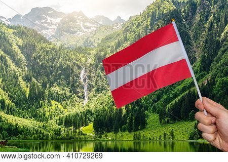 Hand Is Holding Austria Flag In Hand. View On Steirische Bodensee Lake In Austria.