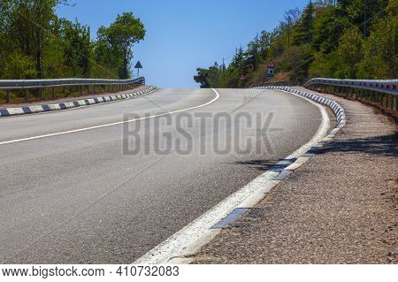 Asphalt Mountain Road With Iron Fence With White Markings. At The Edges Of The Road Are Green Trees