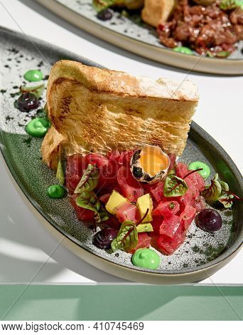 Tuna tartar with quail egg and toast bread. Raw fish tartare restaurant appetizer on white table with green wall. Day sunlight with hard shadow of monstera palm leaves. Summer or spring food concept