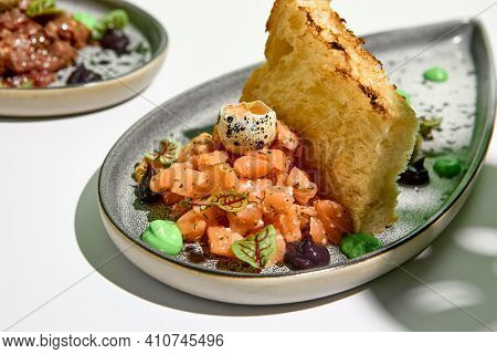 Salmon tartar with quail egg and toast bread. Raw fish tartare restaurant appetizer on white table with green wall. Day sunlight with hard shadow of monstera palm leaves. Summer or spring food concept