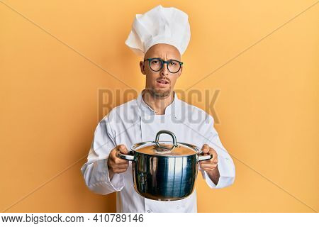 Bald man with beard wearing professional cook holding cooking pot in shock face, looking skeptical and sarcastic, surprised with open mouth 