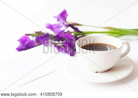 A White Cup With Black Coffee And A Bouquet Of Iris Flowers And A Blank Paper Envelope Are On A Ligh
