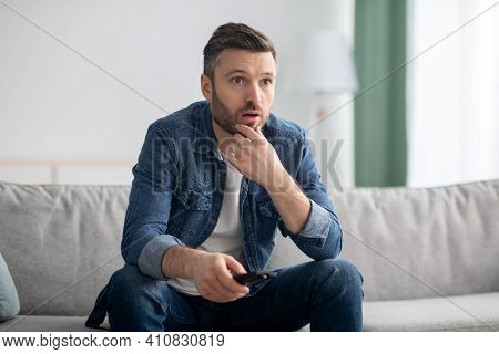 Shocked Middle-aged Beraded Man In Casual Outfit With Tv Remote Sitting On Couch At Home, Watching N