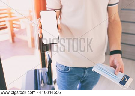 The man holding the mobile phone with empty space as a mockup for digital vaccination certificate in one hand and passport, mask and ticket in another one
