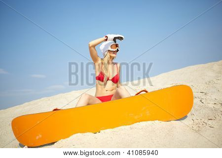 Unga blonda kvinnan sitter på sand med en snowboard i resort omgivningar
