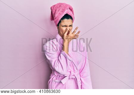 Young hispanic woman wearing shower towel cap and bathrobe smelling something stinky and disgusting, intolerable smell, holding breath with fingers on nose. bad smell 