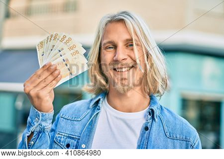 Young blond scandinavian man smiling happy holding danmark 100 kroner banknotes at the city.