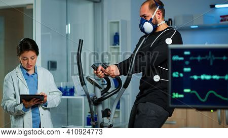 Monitor Showing Ekg Reading Of Athlete Running On Cross Trainer, While Medical Specialists Supervise