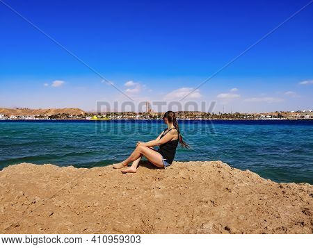 Barefoot Young Woman With Blond Braided Hair Sits On A Rock Against The Background Of The Sea Panora