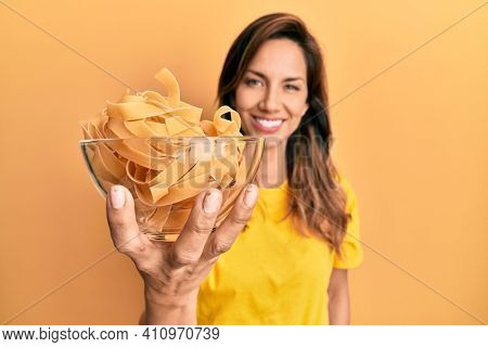 Young latin woman holding bowl with uncooked pasta looking positive and happy standing and smiling with a confident smile showing teeth 