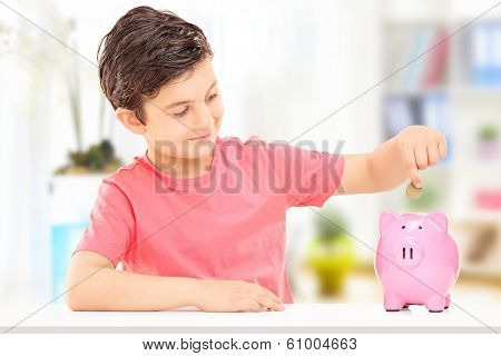 Boy inserting coins into a piggybank seated at table indoors