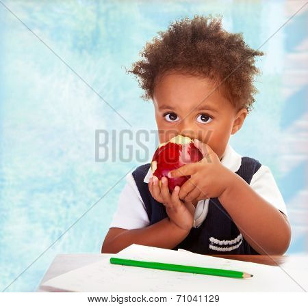 Portrait of cute little African boy sit behind a desk and biting big red apple, drawing using green pencil, having lunch in classroom, happy preschooler concept