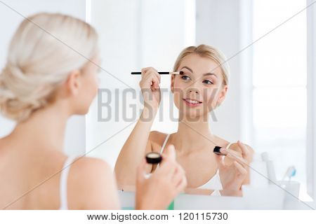 beauty, make up, cosmetics, morning and people concept - young woman applying eyeshade with makeup brush and looking to mirror at home bathroom