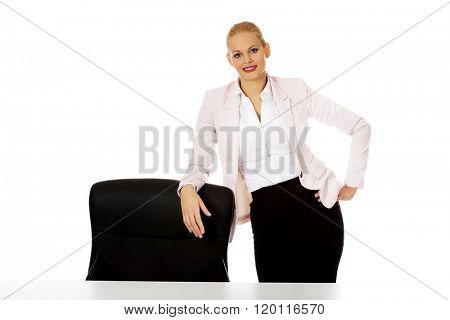 Young busiess woman standing behind the desk leanig on armchair
