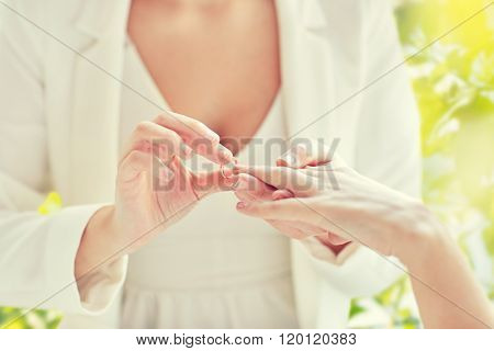 people, homosexuality, same-sex marriage, summer and love concept - close up of happy lesbian couple hands putting on wedding ring over green tree leavers background