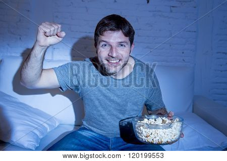 young happy fun sitting at living room couch watching sport match on tv cheering his team gesturing victory fist smiling excited holding popcorn in broadcast television concept