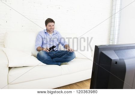 young happy man watching tv lying at home living room sofa with remote control looking relaxed smiling and laughing enjoying television program comedy movie or sitcom