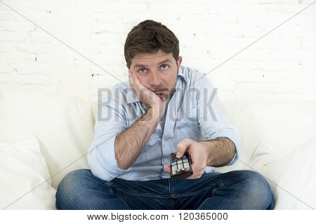 young bored man watching tv sitting at home living room sofa looking tired and not having fun with the television program or movie using remote control for changing to another channel