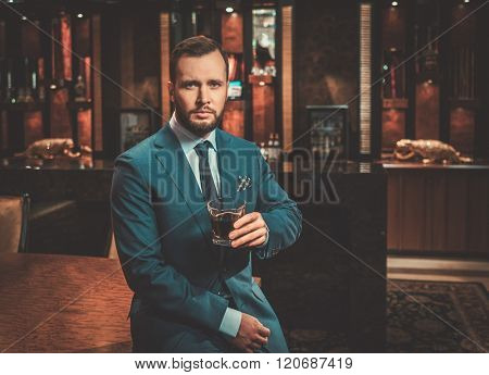 Confident well-dressed man with glass of whisky in luxury apartment interior.