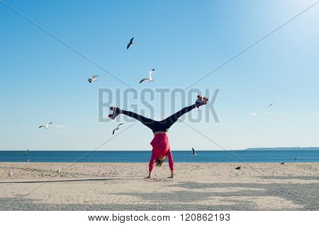Happy young woman doing cartwheel on the beach wearing rollerskates. Portrait of woman on the beach doing a handstand with sea and seagulls in background.
