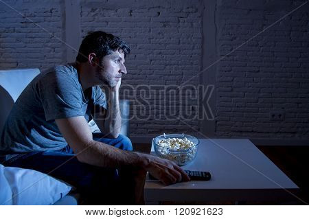 young television addict man sitting on home sofa watching TV eating popcorn using remote control looking bored and tired zapping foe another movie sitcom or live sport at night