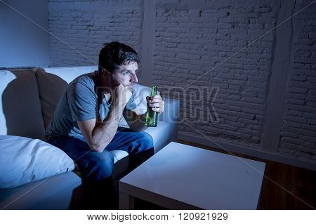 young television addict man sitting on home sofa watching TV and drinking beer bottle looking mesmerized enjoying movie sitcom or live sport at night
