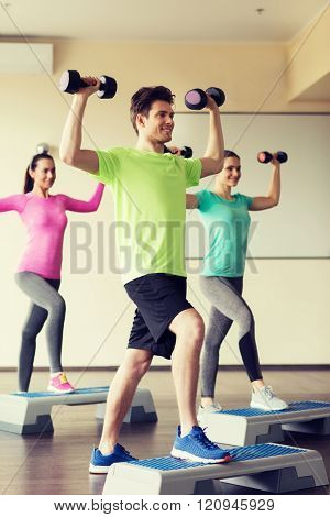 fitness, sport, aerobics and people concept - group of smiling people working out with dumbbells flexing muscles on step platforms in gym