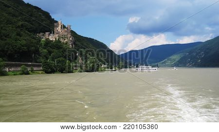 Ancient,castle Rheinstein on the rocky cliff along the river Rhine above the green forest while the white turistic ship sail to the castle to embarkart the guests