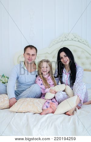 Child with mom and dad sit on bed, smiling and looking at camera. Young happy couple together. Woman with long black hair, brown man and daughter blonde European appearance against white interior. Concept of family life, happiness and joy.