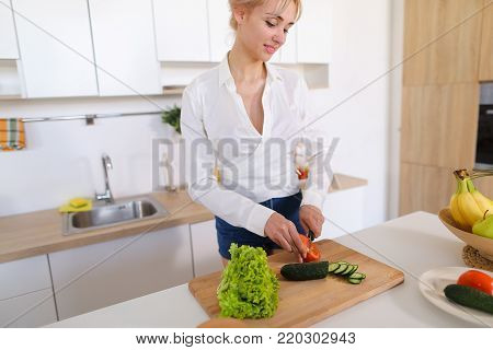 Attractive young woman cuts green cucumber for salad with sharp knife, takes red juicy tomato and several times throws up with smile on face, sniffs freshness of lettuce leaves and takes up shredding vegetables. Blonde at top of modern light kitchen with 