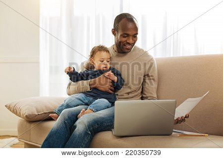 Paternal leave. Cheerful dark-eyed bearded afro-american man smiling and holding his son on his lap while working on the laptop and holding a sheet of paper