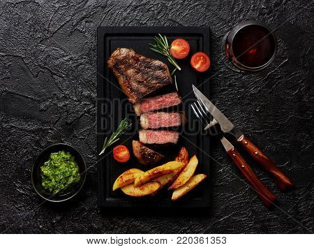 Meat Picanha steak, traditional Brazilian cut with potato wedges, chimichurri sauce and rosemary on black meat cutting board. Steak and a glass of red wine with fork and knife.