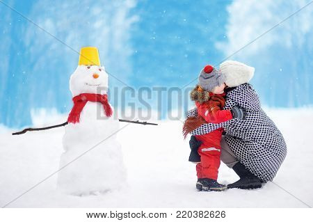 Little boy with his mother/babysitter/grandmother building snowman in snowy park. Active outdoors leisure with children in winter. Kid embrace with his mother