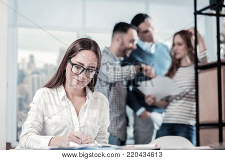 Work with pleasure. Beautiful bespectacled concentrated woman sitting in the office writing and looking down.