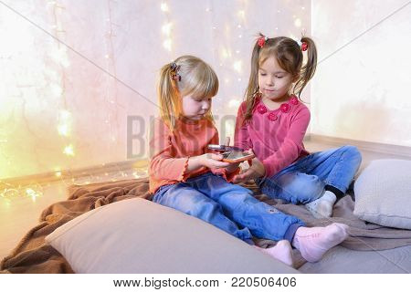 Lovely little girls holding mobile phones and playing games or taking pictures,  typing text for messages on touch screen, they laughing and smiling, sitting on rug in bright room with flickering New Year's garland in evening. Blond girl of European appea