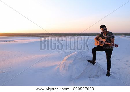 Romantic bearded young Arab man exercises musical instrument with strings, smiles and becomes comfortable in position to start playing guitar, standing in middle of wide sandy desert on warm summer evening at sunset. Swarthy man with dark hair and short 