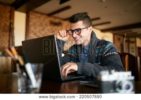 Young attractive hipster in glasses and winter sweater working on laptop in cafe. Freelancer businessman browsing internet at coffeshop. Photographer or designer making project. Vintage camera on table.